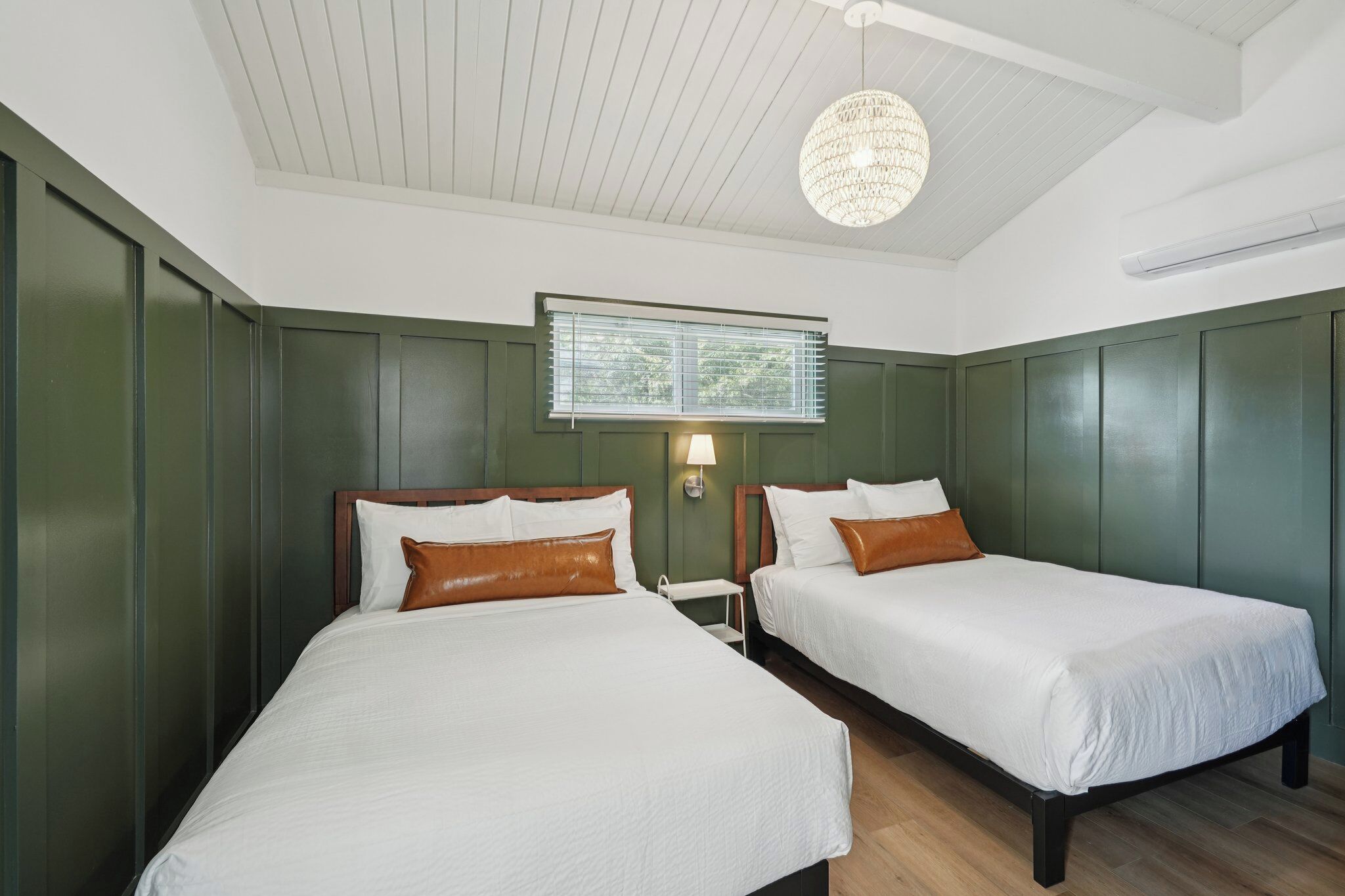 A hotel room with two beds, a chandelier, a window with blinds, and a wall-mounted air conditioner.