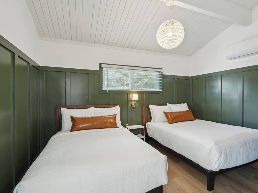 A hotel room with two beds, a chandelier, a window with blinds, and a wall-mounted air conditioner.