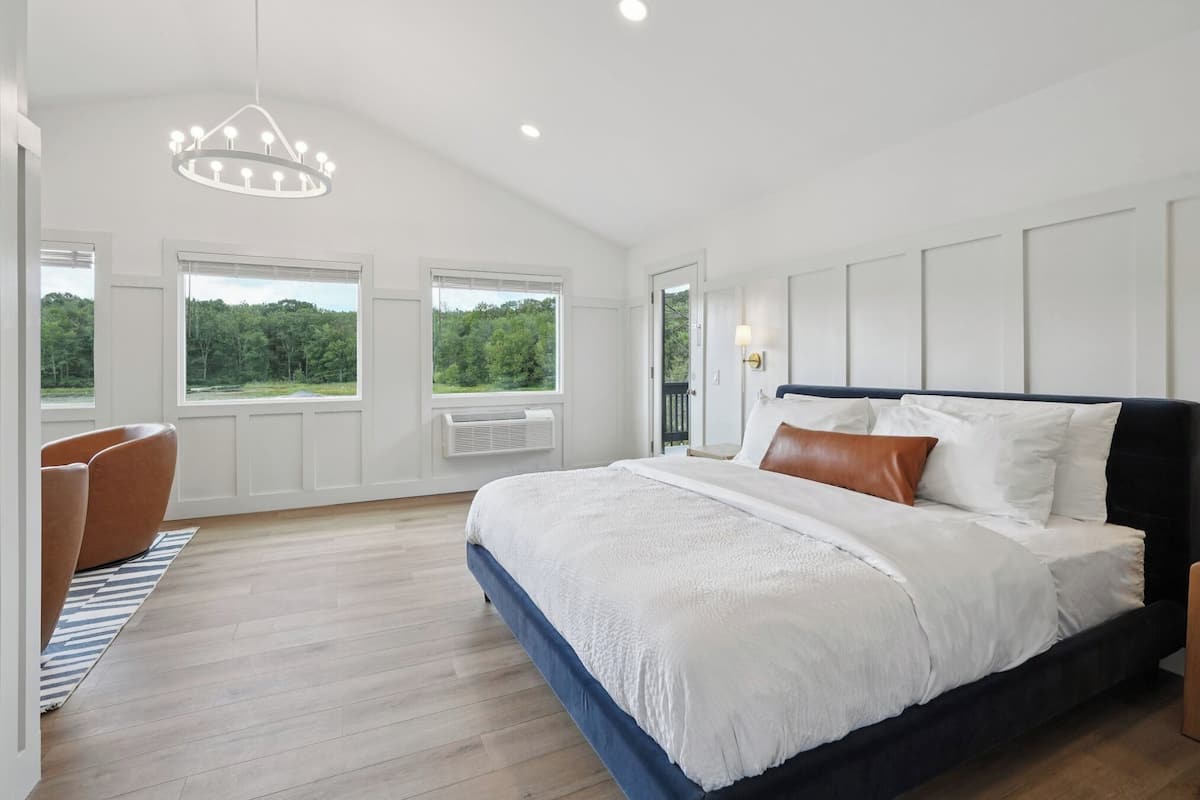 A bedroom with a large bed, a chandelier, a window with a view of trees, and a chair.