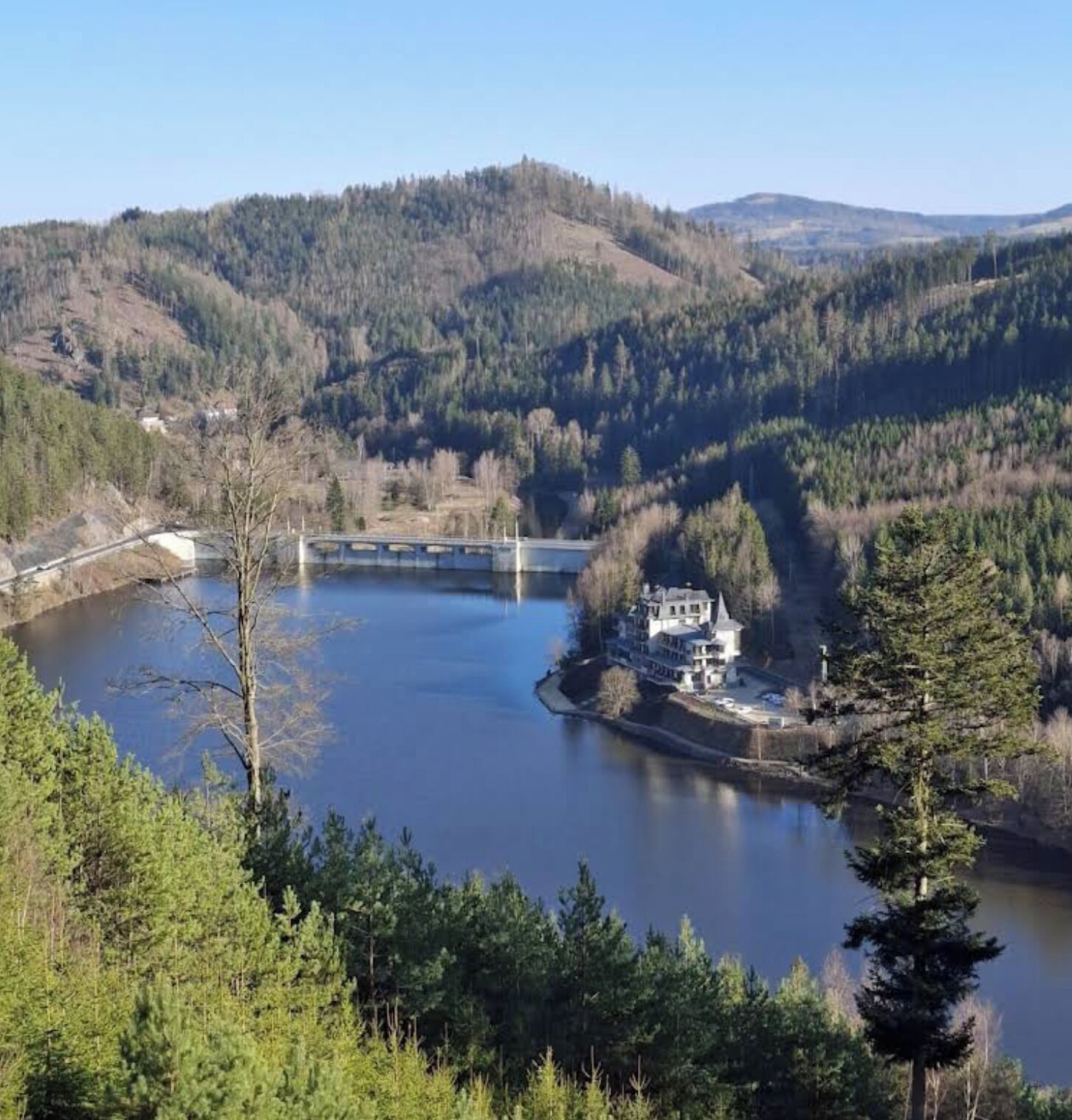 Une vaste étendue d’eau entourée d’une forêt dense et de collines. Un pont enjambe l’eau, et une grande maison est visible sur le côté droit de l’image.
