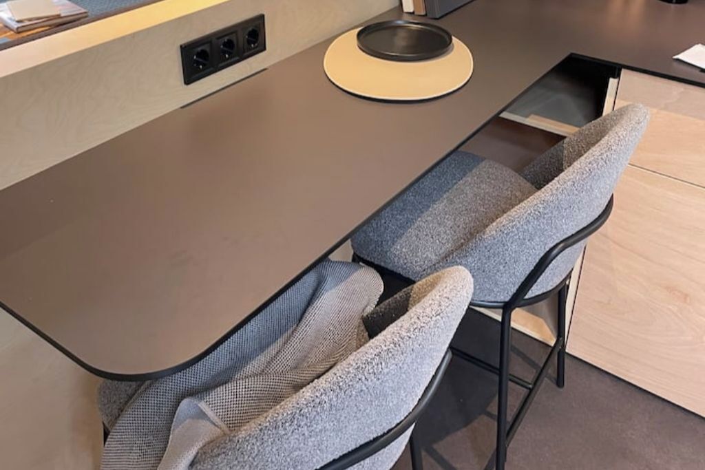 Een moderne keuken met een gebogen aanrecht, twee barkrukken met beklede zittingen, en een inbouwstopcontact.