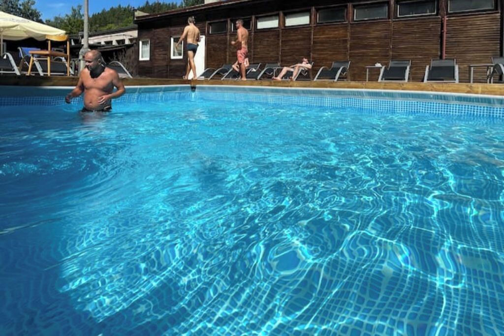 Uma piscina com água azul cristalina, rodeada por espreguiçadeiras e um edifício de madeira ao fundo.