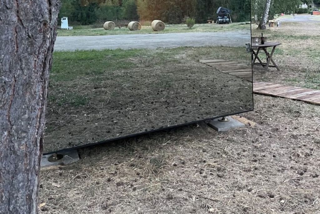En stor, flad, mørk overflade med metalramme, muligvis et bord eller en platform, i et udendørs miljø med et træ, en træterrasse og høballer i baggrunden.