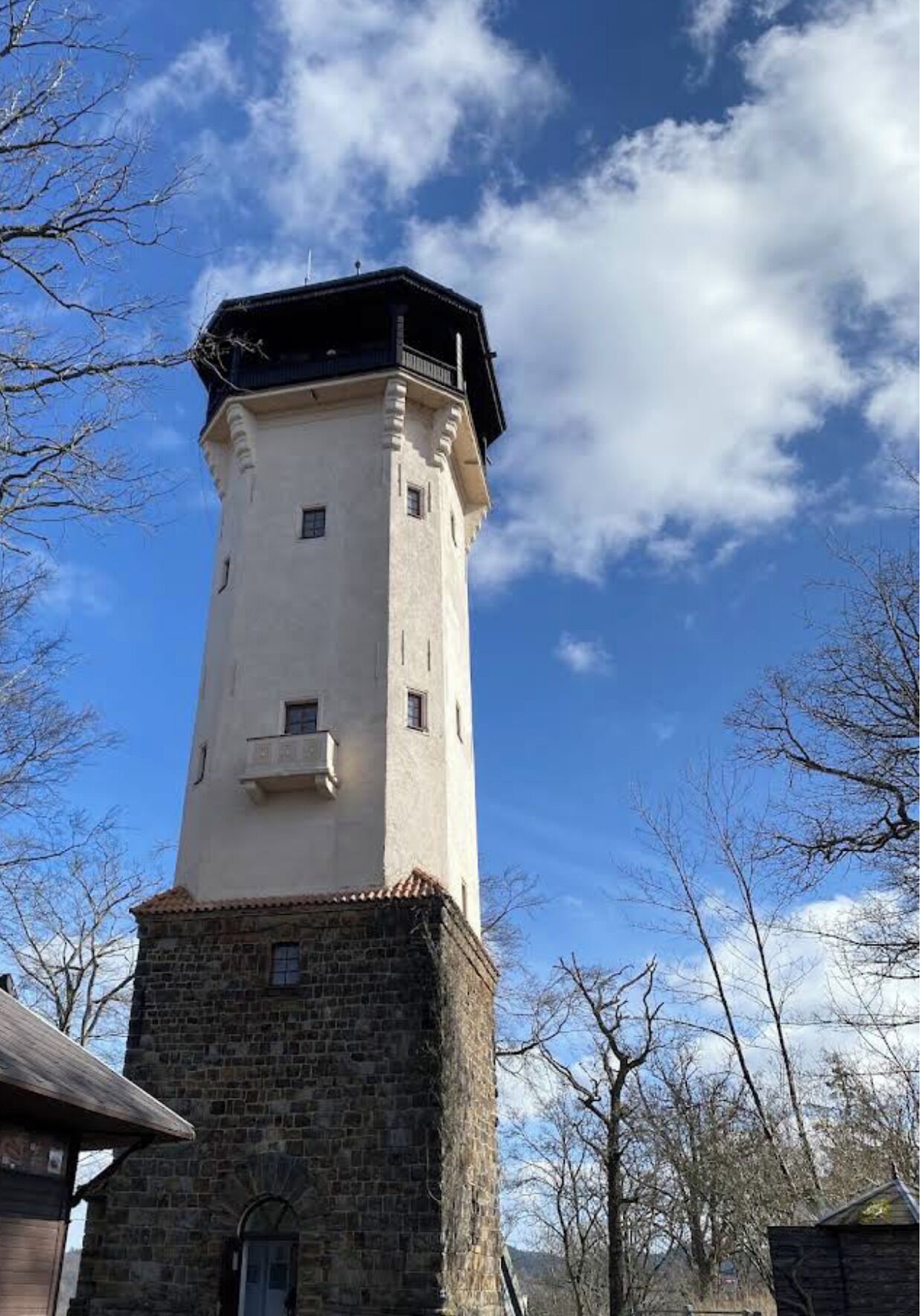 Une haute tour blanche, avec une base en pierre, entourée d’arbres et se dressant sous un ciel dégagé.