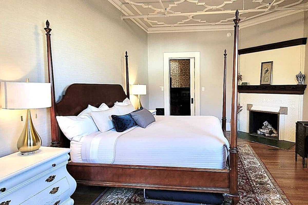 Un lit à baldaquin avec un couvre-lit blanc et de nombreux oreillers, une table de chevet blanche avec une lampe, un bureau en bois avec un téléviseur à écran plat, et une cheminée en arrière-plan.