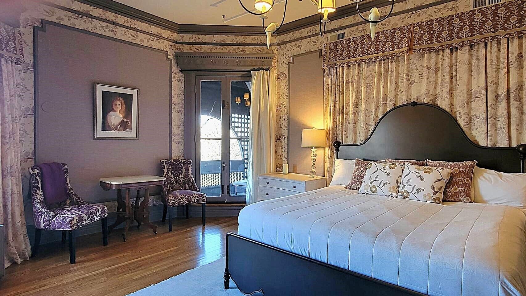 A bedroom with a large bed, a desk, two chairs, and a window with curtains.