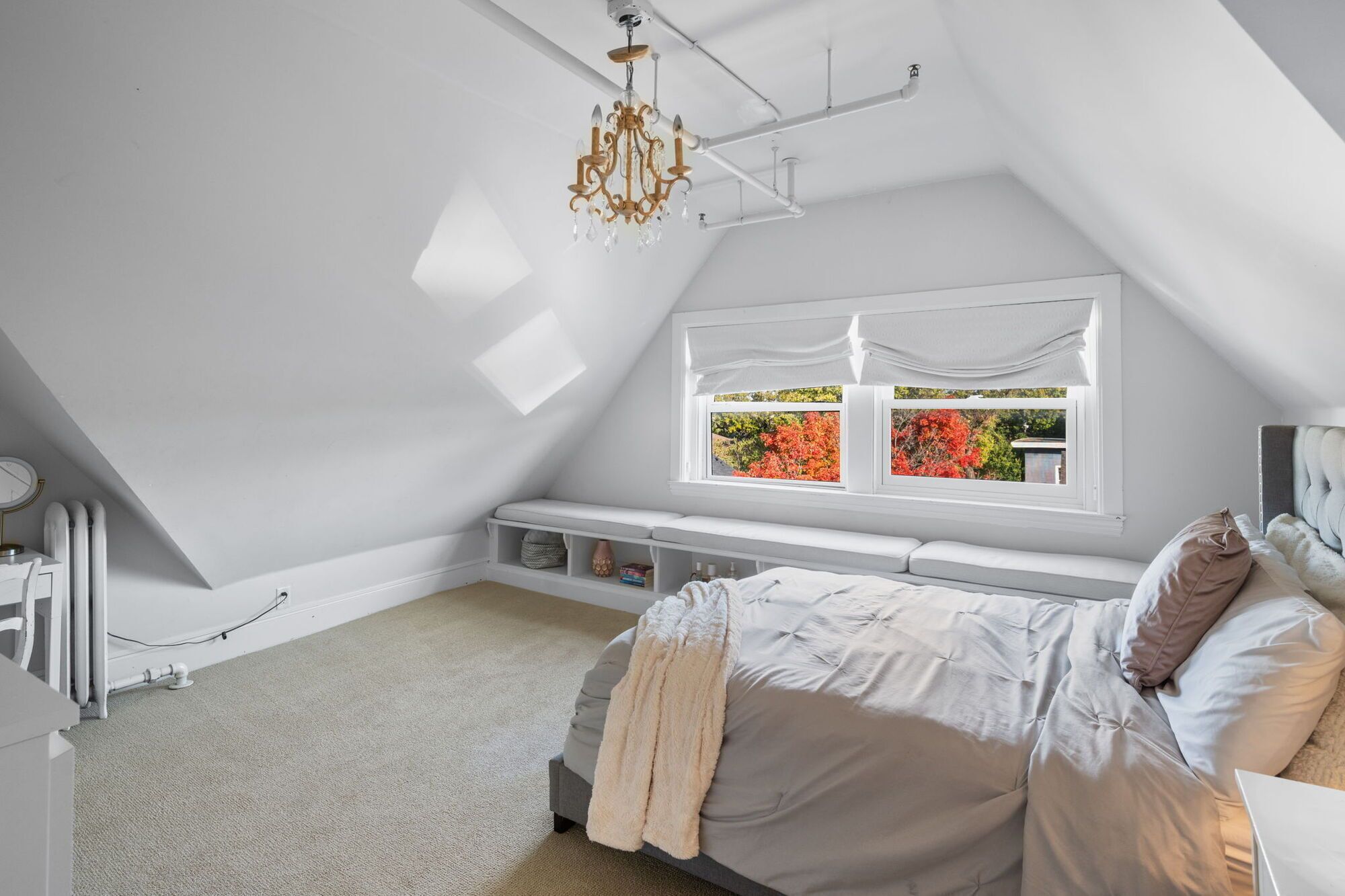 Quarto com teto inclinado, cama com manta bege, mesa de cabeceira, janela com vista para folhagem de outono e um lustre.