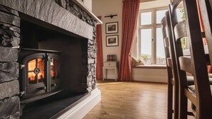 A cozy room with a stone fireplace, wooden flooring, a window with red curtains, and a white wall with framed pictures. - 2 Hart Head Barn, Beautiful Renovated Barn in Rydal Sleeping 10. (Ambleside)