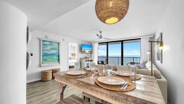 Un coin repas avec une table en bois dressée pour quatre personnes, un ventilateur de plafond et une grande fenêtre donnant sur l’océan.