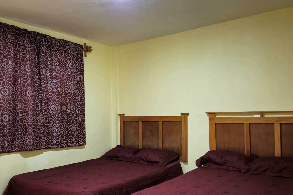Une chambre avec deux lits simples, une tête de lit en bois, un rideau à motifs et un sol carrelé.