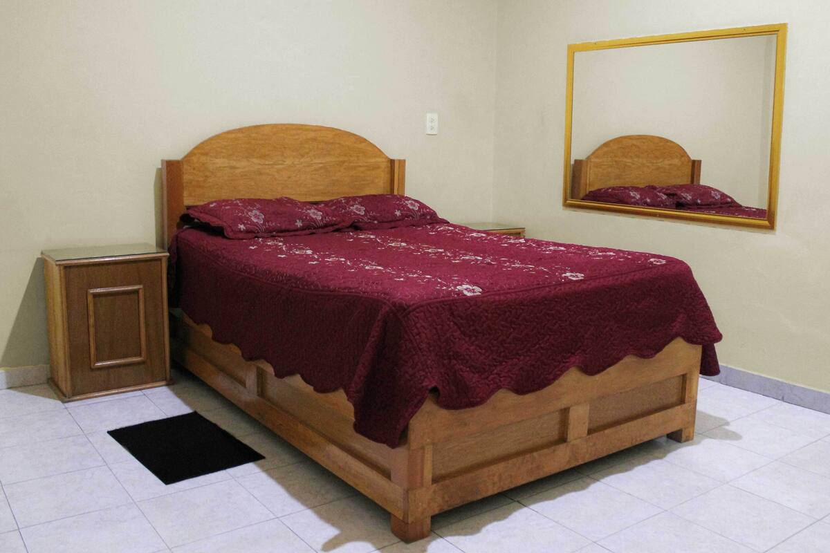 Un lit simple avec une tête de lit en bois et une table de chevet assortie, un miroir au mur et un tapis noir sur le sol.