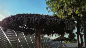 A round table with a thatched roof, surrounded by stone stools, set on a gravel surface with a fence and trees in the background. - Hotel San Jose  (Tepeaca)