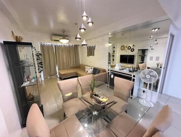 A modern living room with a glass dining table, beige sofas, and a kitchen area visible through a glass partition.