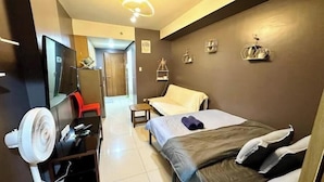 A hotel room with a bed, a desk with a chair, a TV, a fan, and a door leading to another room. - Kathy’s Crib Family Suite Room at Shell Residence (Pasay City)