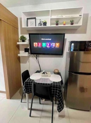 A compact kitchen with a flat-screen TV mounted on the wall, a refrigerator, and a dining table with chairs.