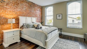 Une chambre avec un mur en briques, un lit blanc, un ventilateur de plafond et de grandes fenêtres.