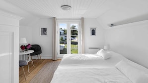 Une chambre à coucher blanche avec un lit, une chaise, une petite table ornée de fleurs et une vue sur l’extérieur, visible à travers une porte.