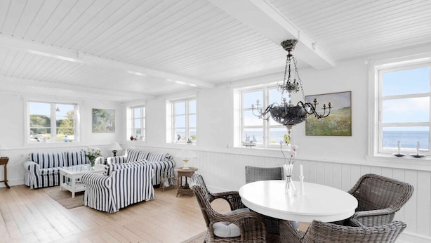 Une pièce spacieuse avec un plafond haut, des murs blancs et de grandes fenêtres. On y trouve une table à manger ronde, entourée de chaises en osier, et un lustre. Un ensemble de canapés à rayures est placé contre le mur, et une chaise en osier est disposée devant la table.