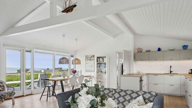 A spacious living area with a large window overlooking a scenic view, a dining area with a table and chairs, and a kitchen with white cabinets and a wooden countertop. - 5 Star Holiday Home in Bogense (Bogense)
