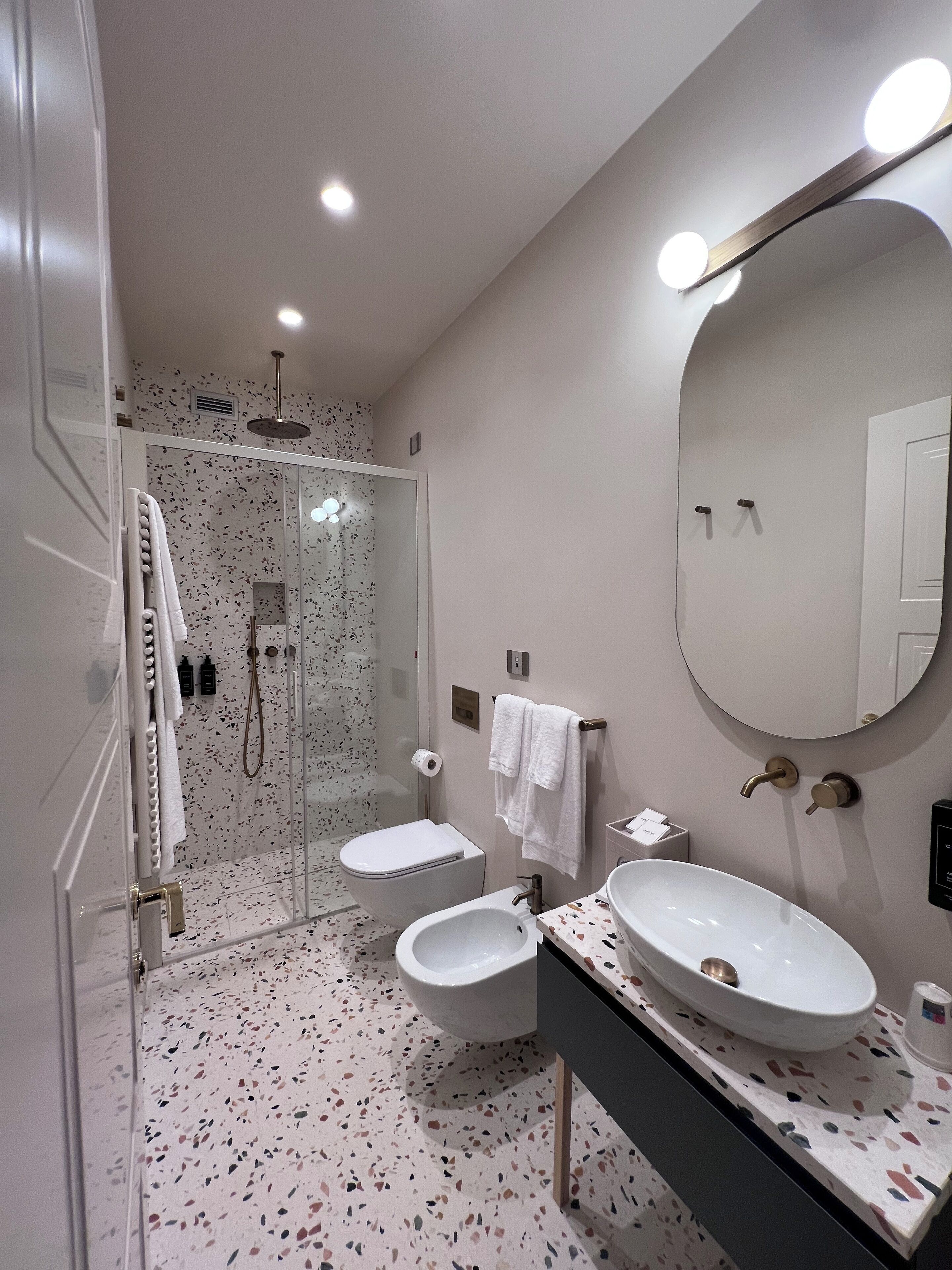 Un baño moderno con ducha de vidrio, dos lavamanos, un espejo grande y un suelo moteado.