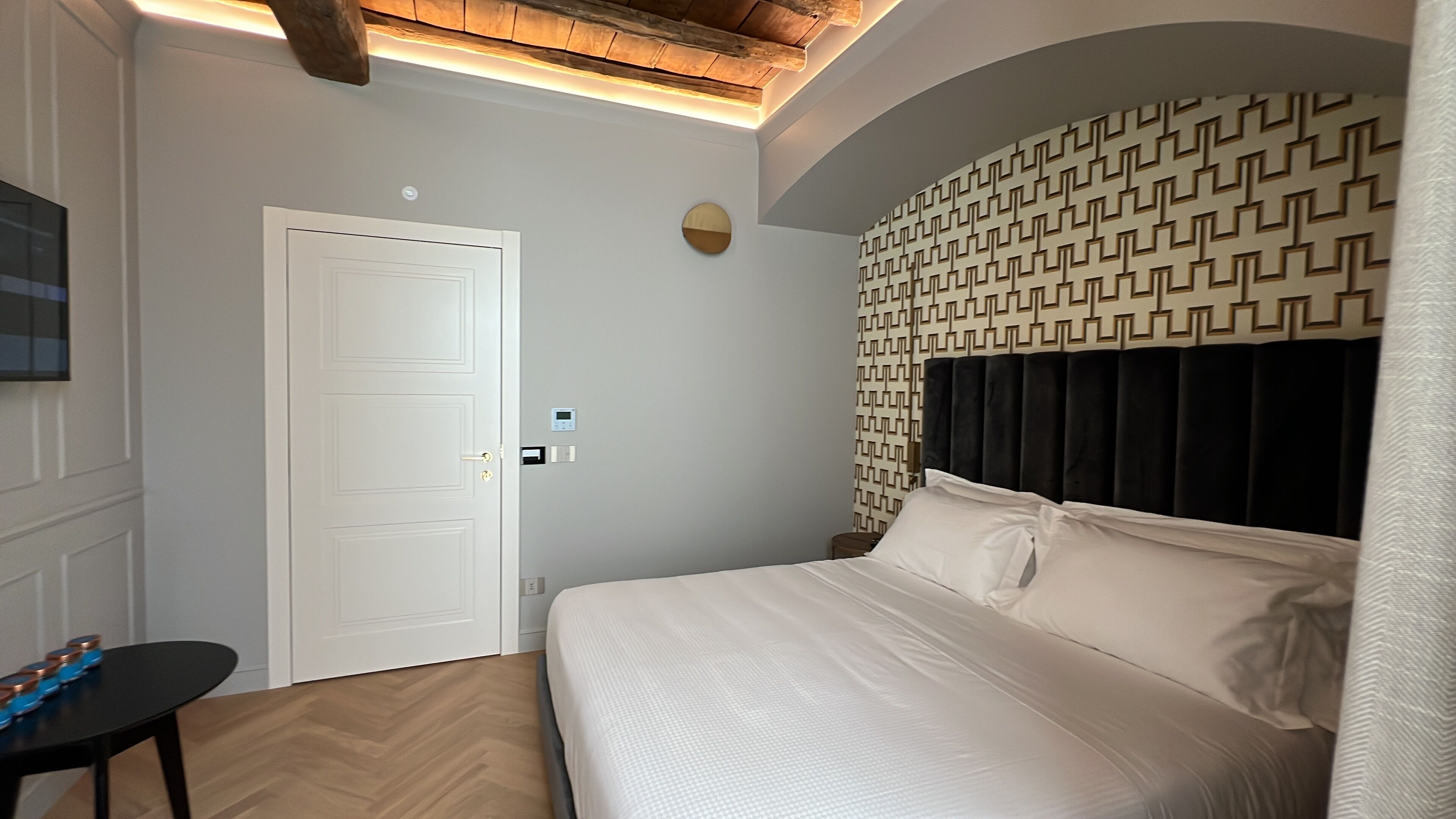 A bedroom with a bed, a white door, a small table, and a patterned wall.