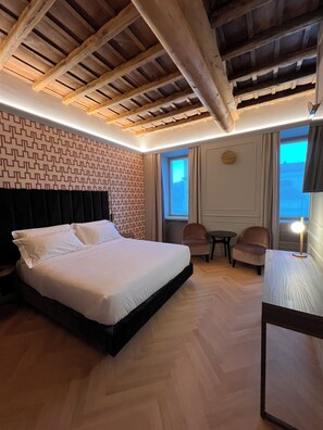 Une chambre d’hôtel avec un grand lit, des poutres en bois au plafond, un mur décoré d’un motif, une petite table, deux chaises et une fenêtre avec des rideaux.