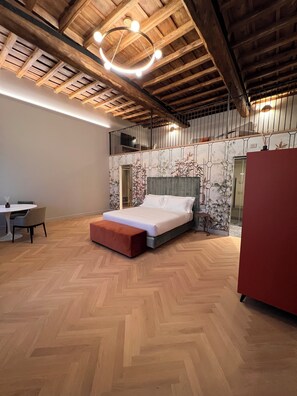 Une chambre avec un plafond en bois, un lit doté d’une tête de lit grise, un banc rouge et un bureau avec des chaises.