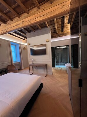 A modern hotel room with a wooden ceiling, a large bed, a TV mounted on the wall, and a glass door leading to a bathroom. - Villa Clementi Boutique Hotel (Cave)