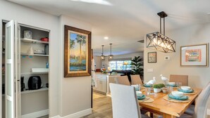 A dining area with a wooden table set for four, a kitchen with open shelving, and a painting on the wall. - Waterfront Home w/ Boat Dock & Bar in N Fort Myers (North Fort Myers)