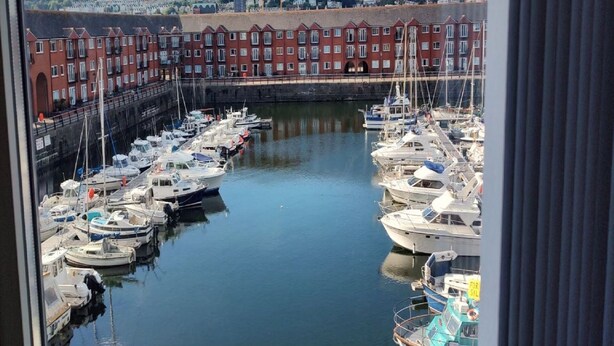 A marina with multiple boats docked, surrounded by red brick buildings and a hilly landscape in the background. - Marina View Apartment (Swansea)