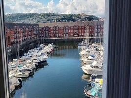 A marina with multiple boats docked, surrounded by red brick buildings and a hilly landscape in the background.
