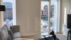 A living room with a sofa, a glass coffee table, and a TV. Large windows offer views of a marina and buildings. - Marina View Apartment (Swansea)