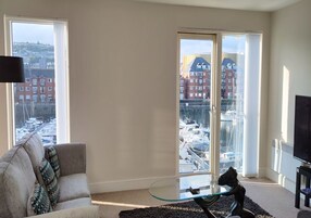 A living room with a sofa, a glass coffee table, and a TV. Large windows offer views of a marina and buildings.