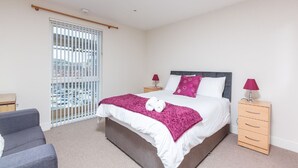 A bedroom with a bed, two bedside tables, a chair, and a window with blinds. - Marina View Apartment (Swansea)
