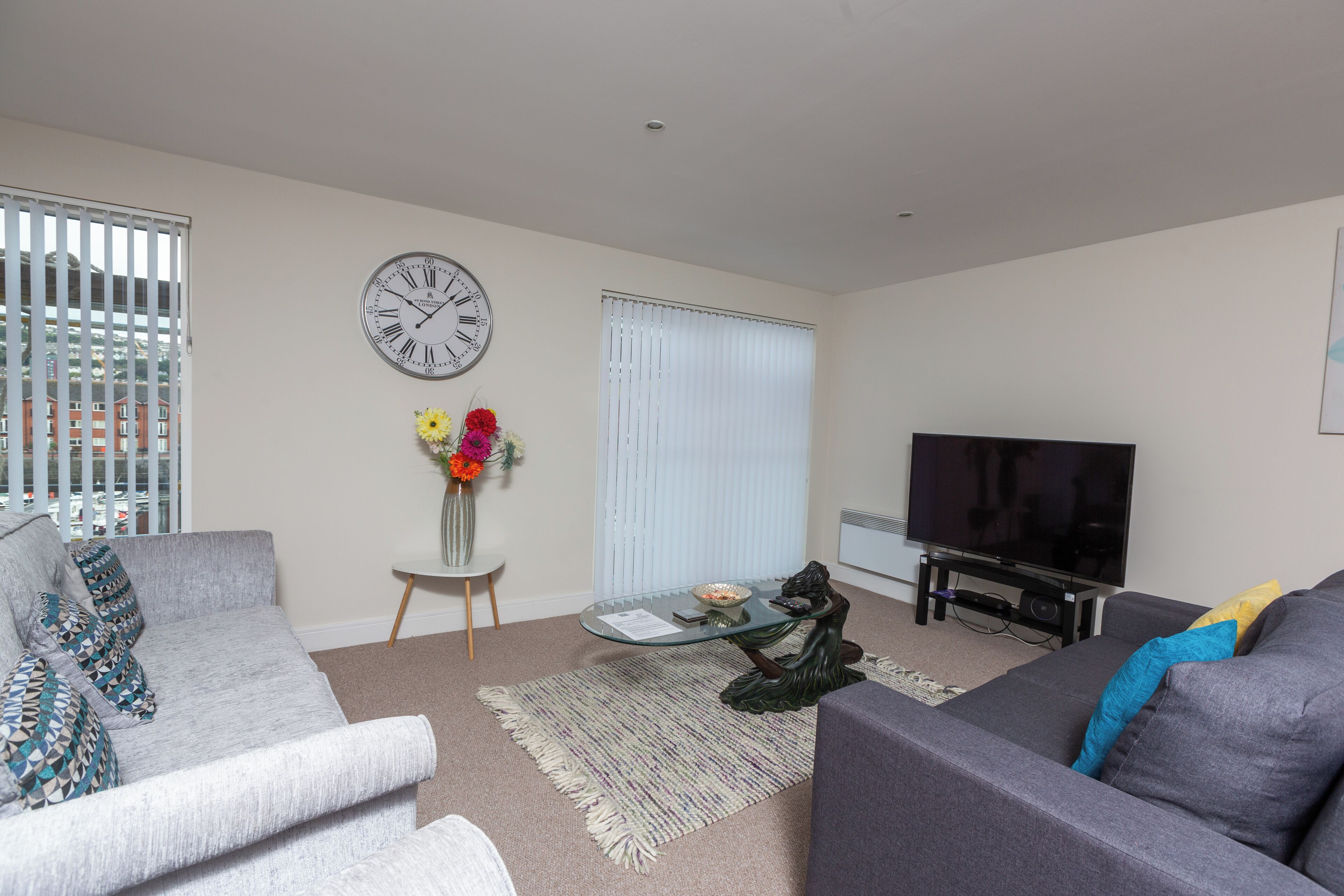 A living room with a grey sofa, a glass coffee table, a TV, a clock, and a window with blinds.