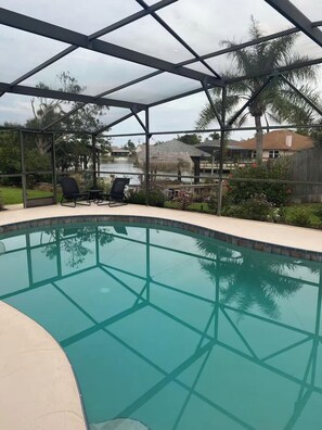 A clear swimming pool with a glass enclosure, surrounded by lounge chairs and a view of a residential area with houses and palm trees.