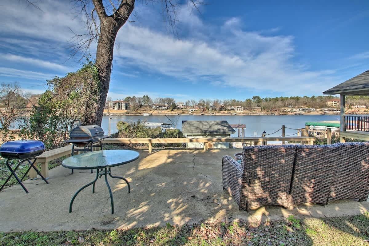 Une terrasse extérieure avec une table et des chaises, un barbecue et une vue sur un lac, avec des bâtiments en arrière-plan.