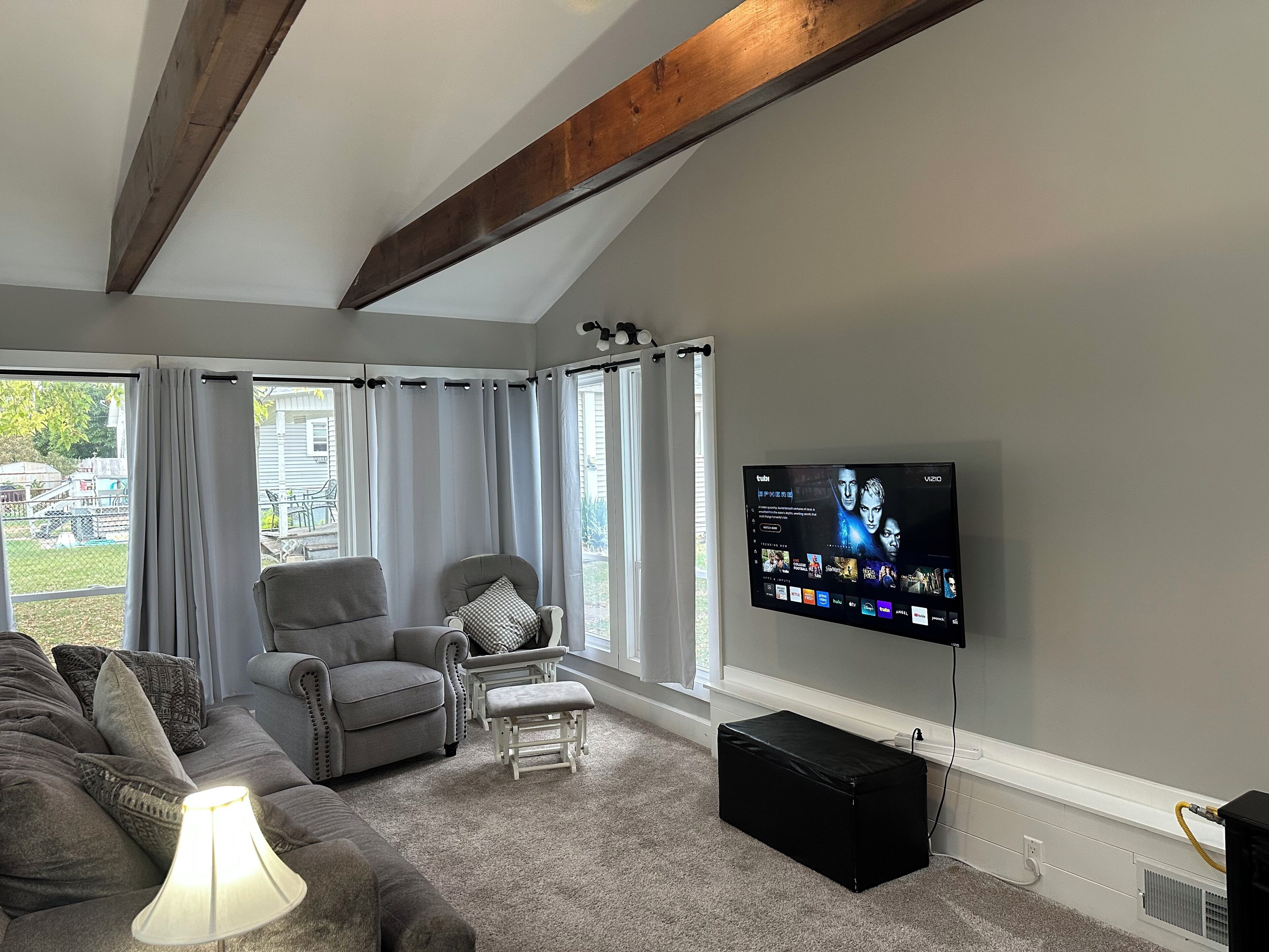 Un salon avec un canapé gris, un fauteuil blanc, une petite table, un pouf noir et un téléviseur à écran plat fixé au mur.