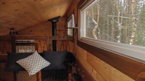 Een gezellig houten interieur met een houten plafond, een raam met uitzicht op een besneeuwd bos, een metalen grilltafel en een comfortabele bank met gekleurde kussens.