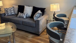 Ein modernes Wohnzimmer mit einem grauen Sofa, gemusterten Kissen, einem Glastisch und zwei schwarzen Stühlen.