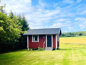 A small red wooden cabin with a tiled roof, situated in a grassy field with trees in the background. - 4 Star Holiday Home in Bralanda (Bralanda)
