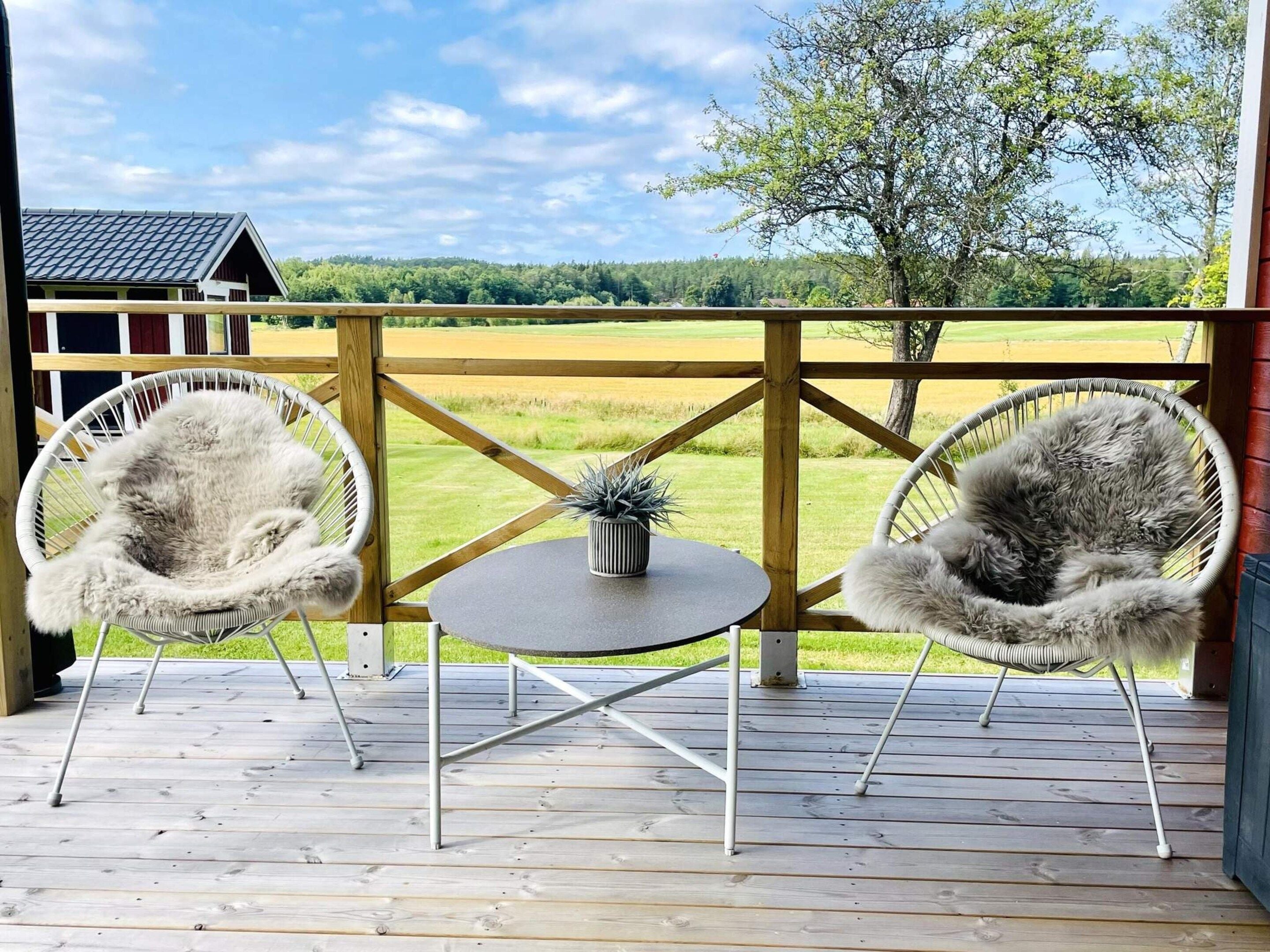 Une terrasse en bois avec deux chaises en osier blanc et une petite table, d’où l’on peut admirer un champ verdoyant et un bâtiment situé au loin.