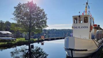 A boat named 'DALSLANDIA' docked at a calm water body with a clear sky and a large tree in the background.
