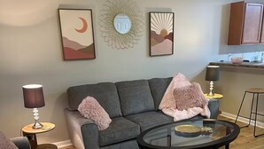Ein Wohnzimmer mit grauem Sofa, einem Glastisch und einer Küchenzeile im Hintergrund.