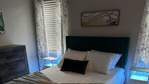 Ein ordentlich bezogenes Bett mit gemustertem Überwurf und Kissen, ein dunkles Kopfteil, ein Nachttisch mit Lampe sowie ein Wandbild.