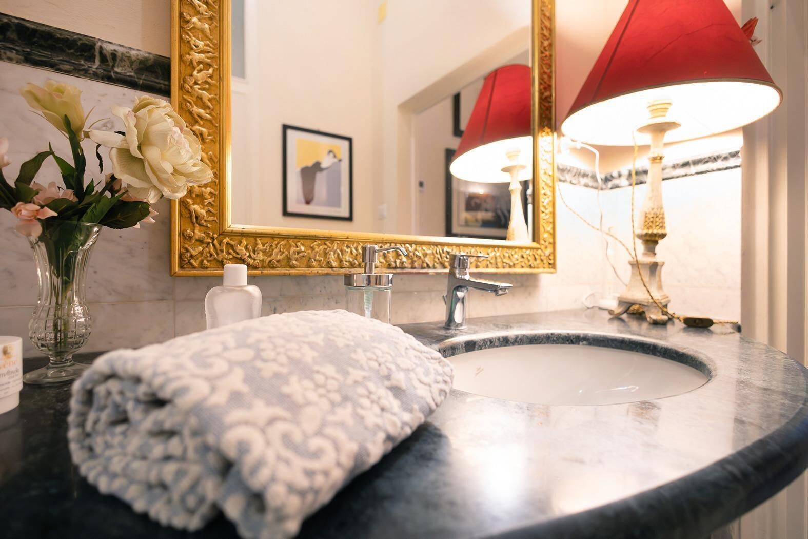 A bathroom with a marble sink, a gold-framed mirror, a red lamp, and a vase with flowers.