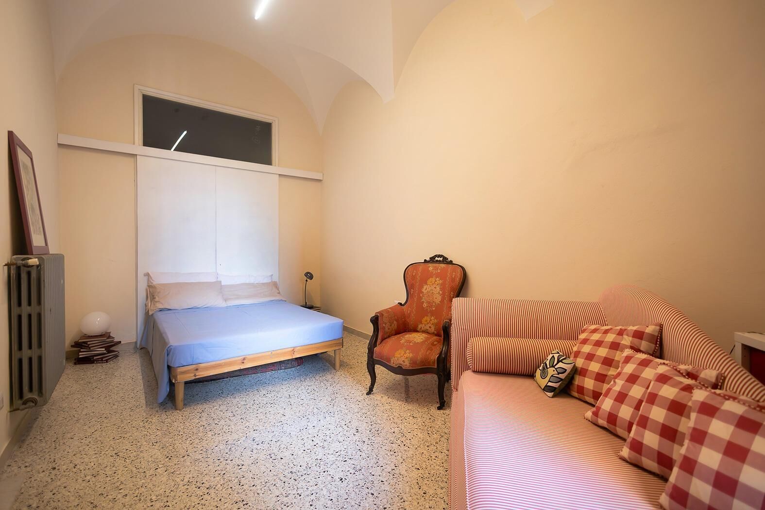 A bedroom with a bed, a chair, a sofa, a radiator, and a window.