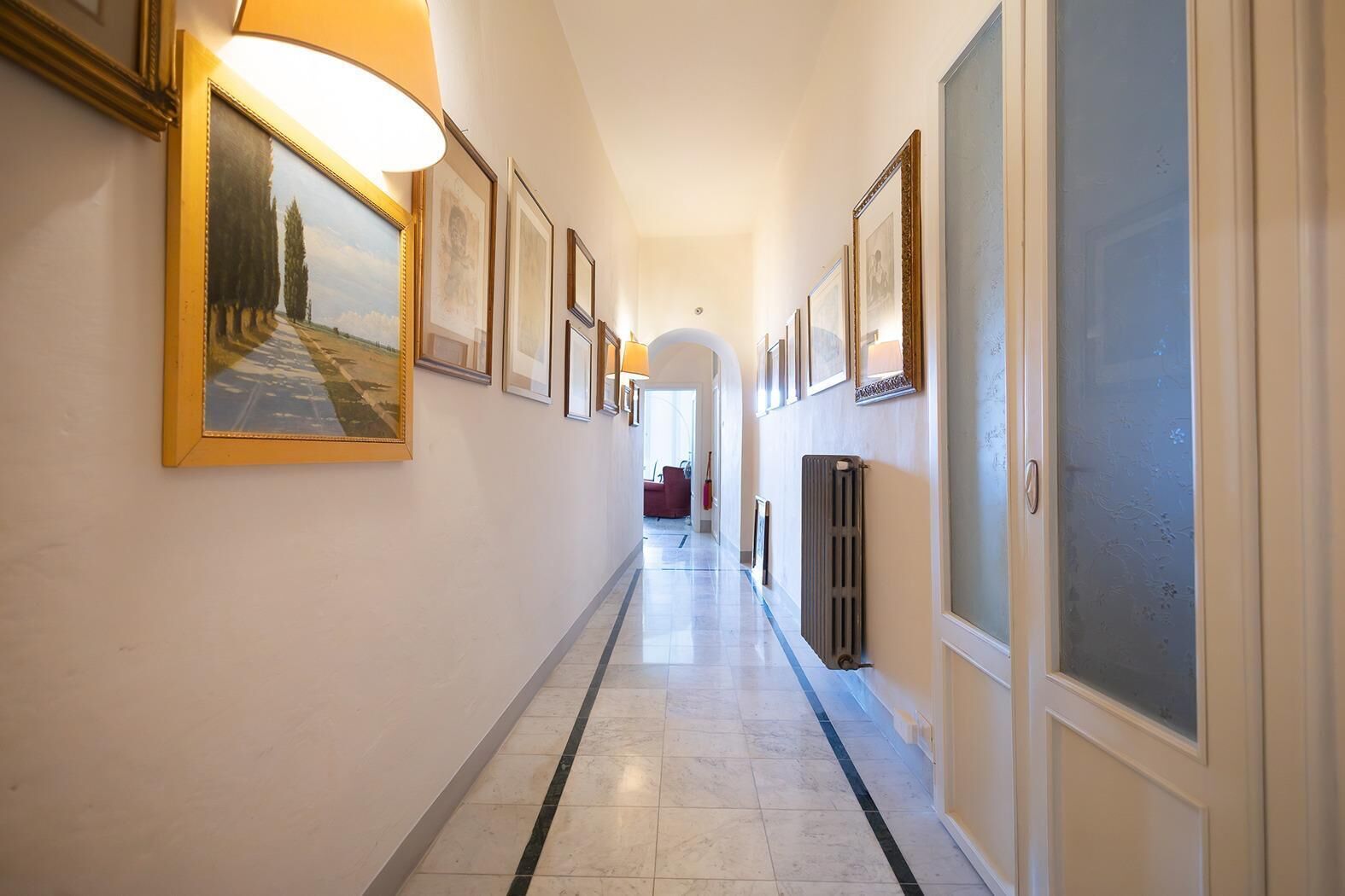A hallway with framed artwork on the walls, a radiator, and a door with frosted glass.