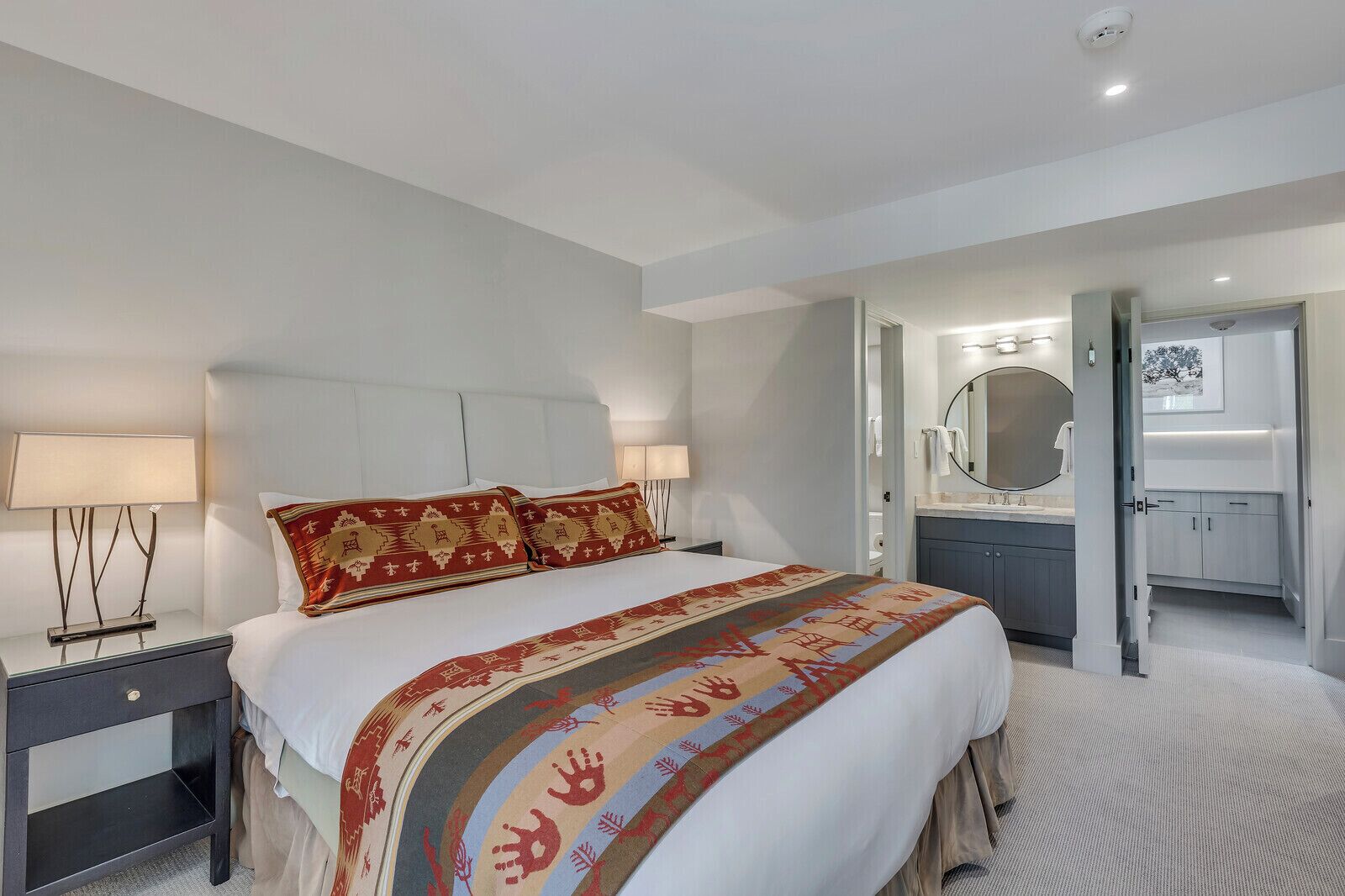 A modern hotel room with a large bed, a nightstand, a bedside lamp, a bathroom with a mirror, and a door leading to another room.