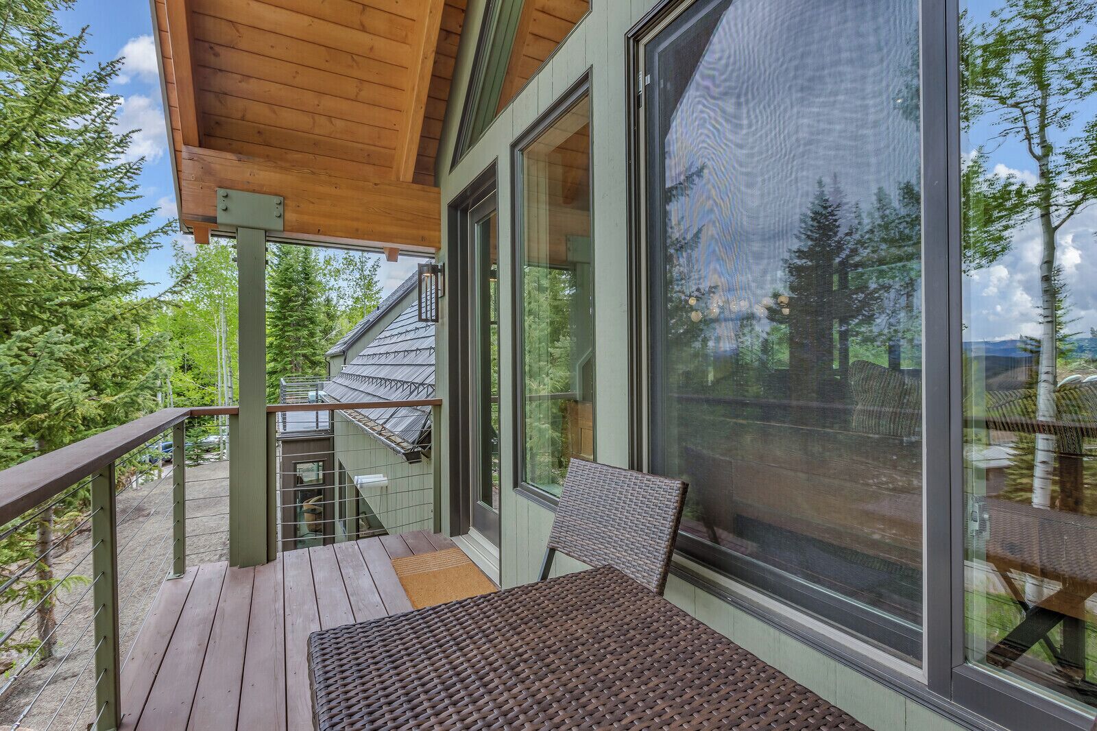 A wooden deck with a table and chairs, large windows offering a view of trees and a distant mountain.